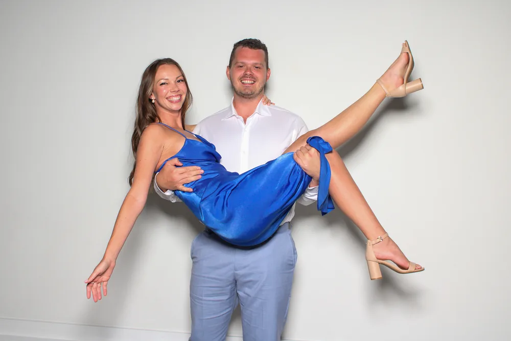 Wedding guest carrying his partner in a blue dress at the photo booth, white backdrop